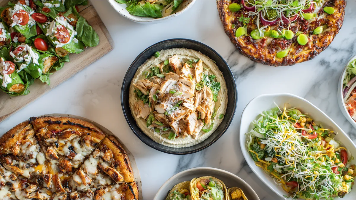 Overhead view of assorted dishes including pizza, hummus with grilled chicken, tacos, and fresh salads on a marble surface